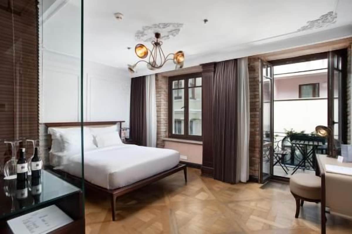 Habitación de hotel con cama, una mesita con botellas, una silla, un balcón con mesa y sillas, y vistas a edificios.