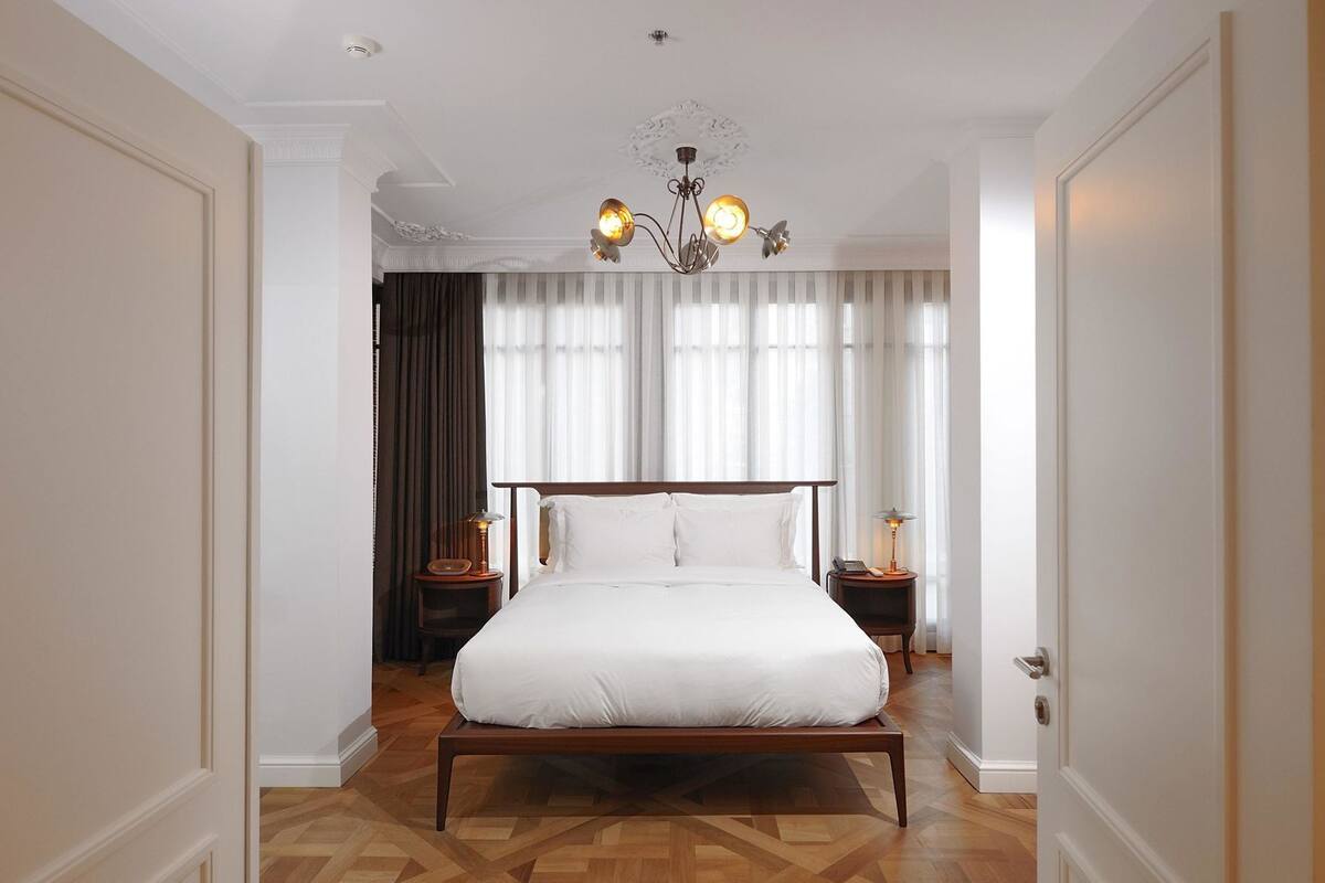 Un dormitorio con una cama grande, dos mesitas de noche, una araña y vistas al exterior a través de cortinas translúcidas.