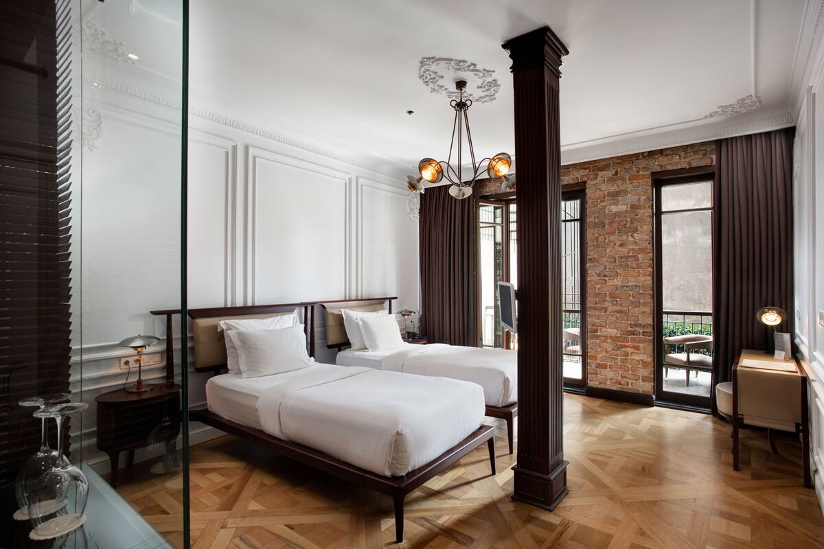 A hotel room with two beds, a chandelier, a brick wall, and a balcony view.