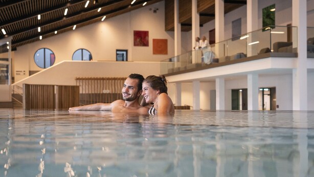 An indoor swimming pool with a modern architectural design, featuring a high ceiling with linear lighting and a glass balustrade overlooking a lower level. - Hotel Summerhof (Bad Griesbach im Rottal)