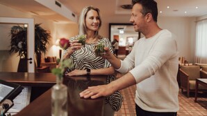 A man and a woman standing at a bar in a hotel lobby with a modern interior design, including a wooden reception desk and a visible menu. - Hotel Summerhof (Bad Griesbach im Rottal)