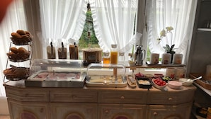 Buffet de café da manhã com diversos alimentos, uma máquina de café e uma árvore de Natal decorativa visível por cortinas translúcidas.