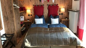 Quarto com parede de madeira, cama com colcha cinza, duas mesas de cabeceira com luminárias, cortina xadrez vermelha e branca, e prateleira de madeira com objetos.