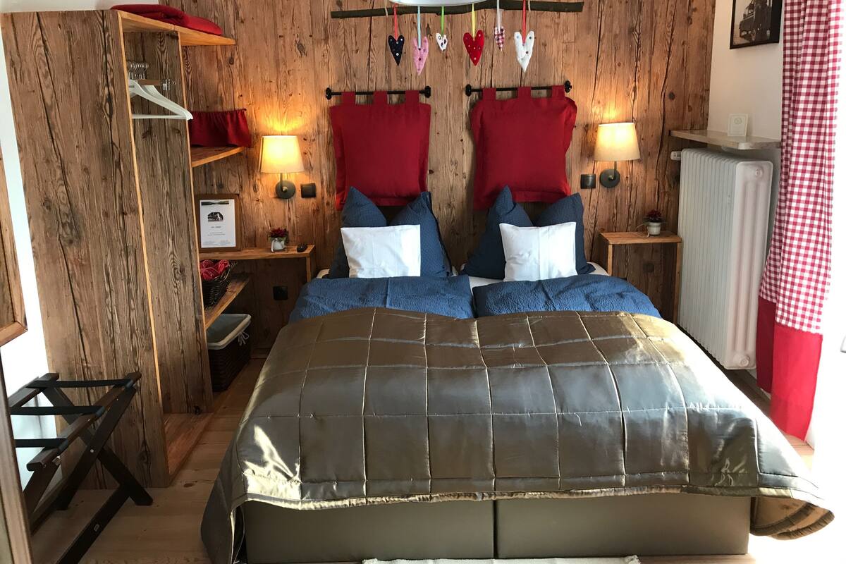 A bedroom with a wooden wall, a bed with a grey quilt, two bedside tables with lamps, a red and white checkered curtain, and a wooden shelf with items.