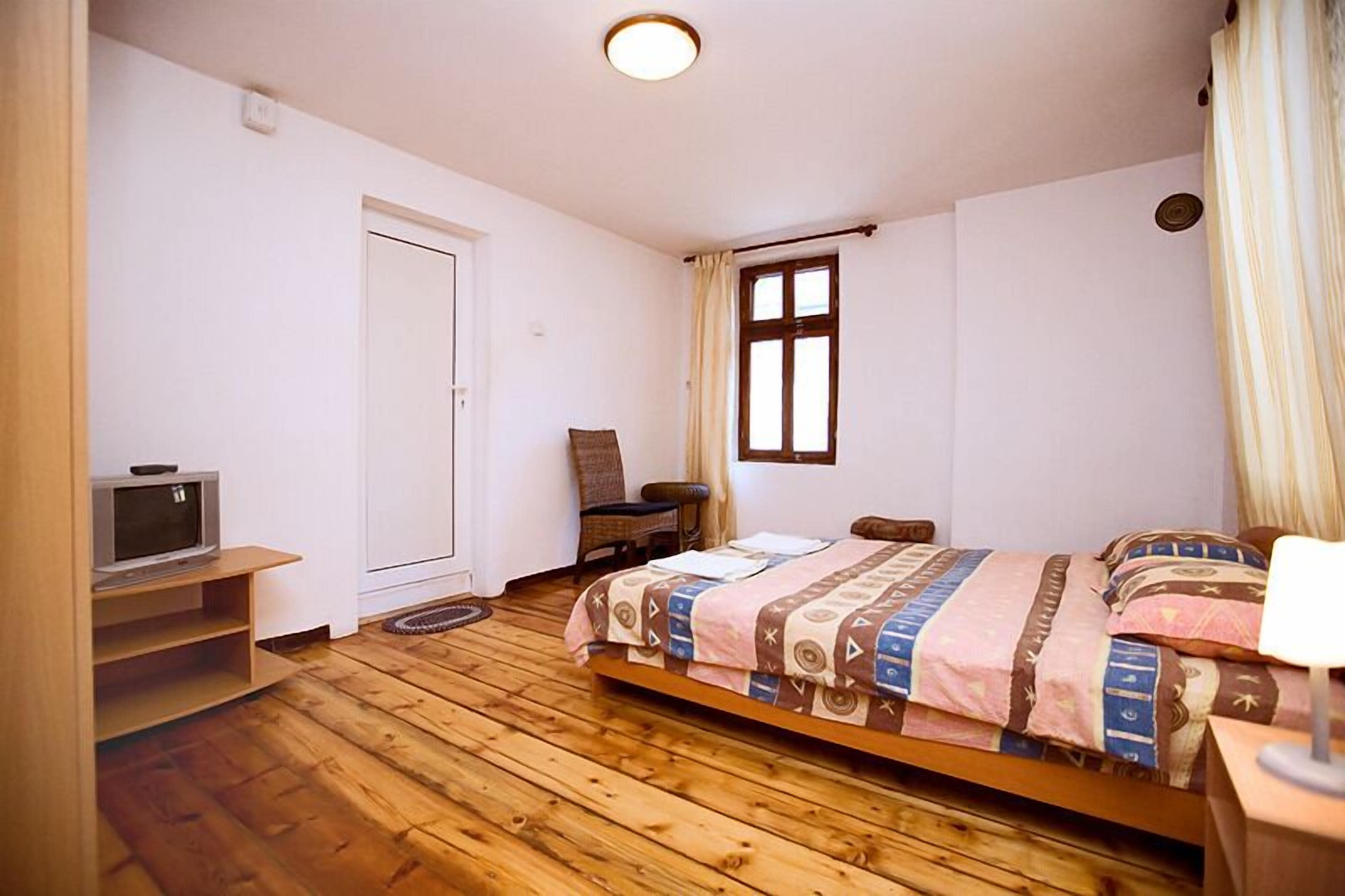 A bedroom with a wooden floor, a bed with a patterned bedspread, a chair, a television on a shelf, and a window with curtains.