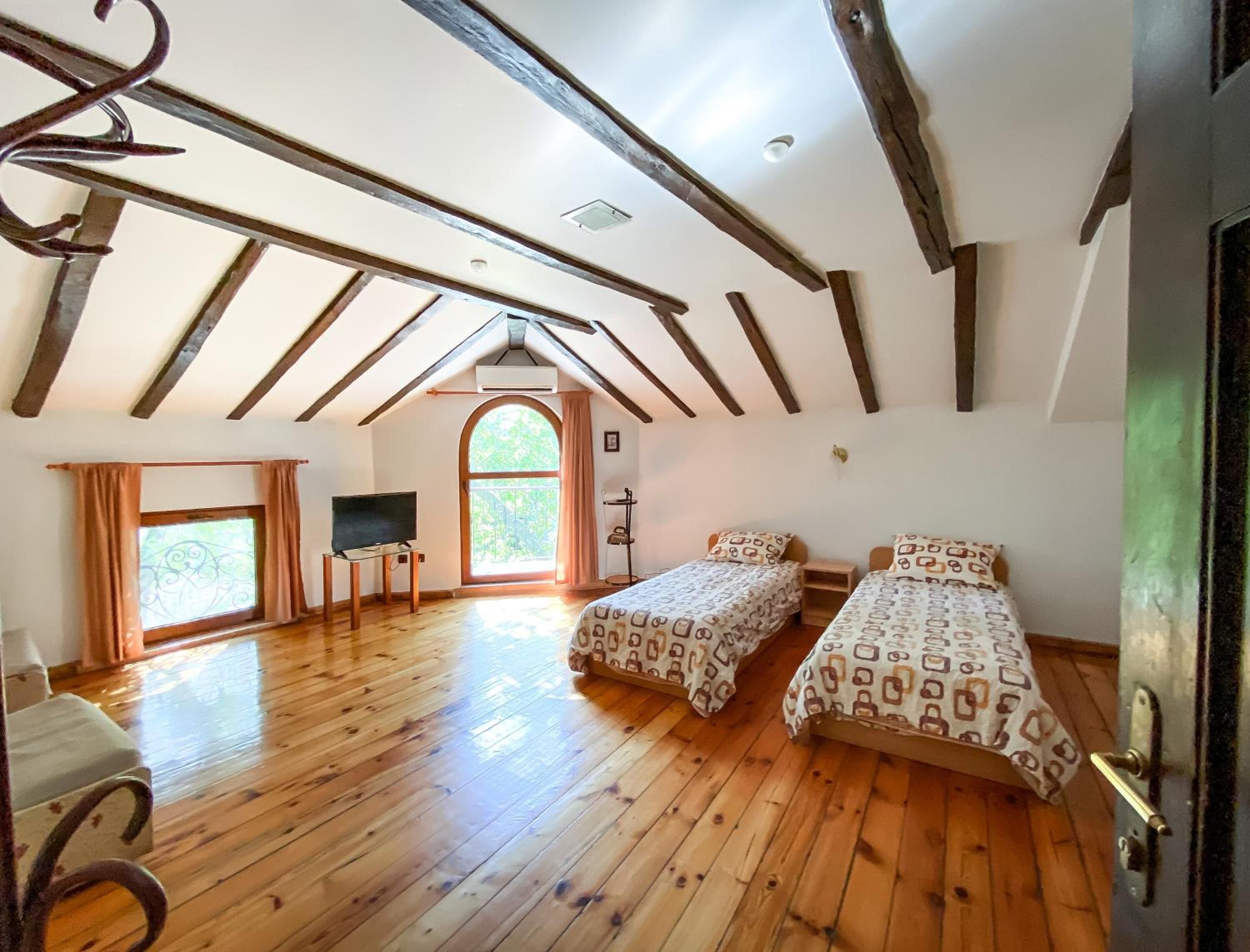 A room with wooden floors, two beds with patterned bedding, a TV, a window with curtains, and a door.