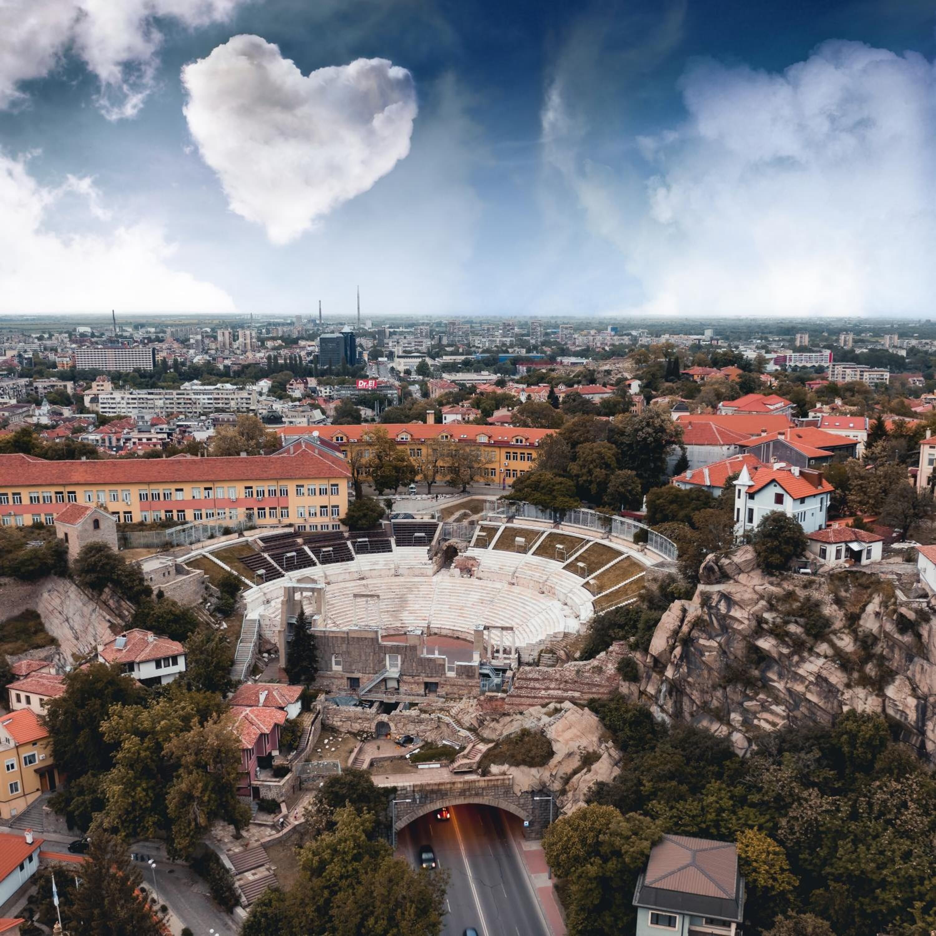 An ancient amphitheater with stone seating and a central stage, surrounded by modern buildings and a road.