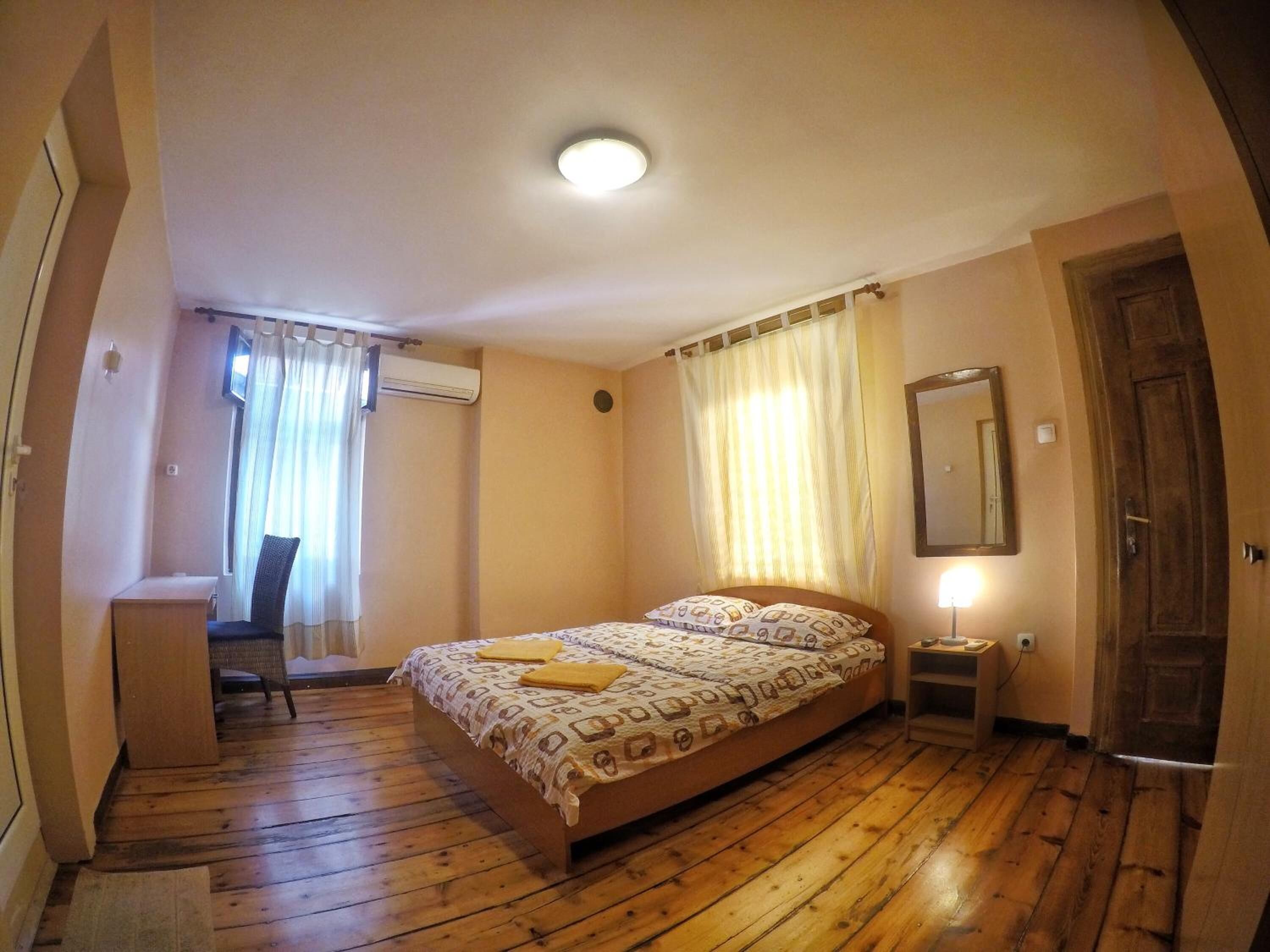 A bedroom with a wooden floor, a bed with patterned bedding, a desk, a chair, and a mirror on the wall.