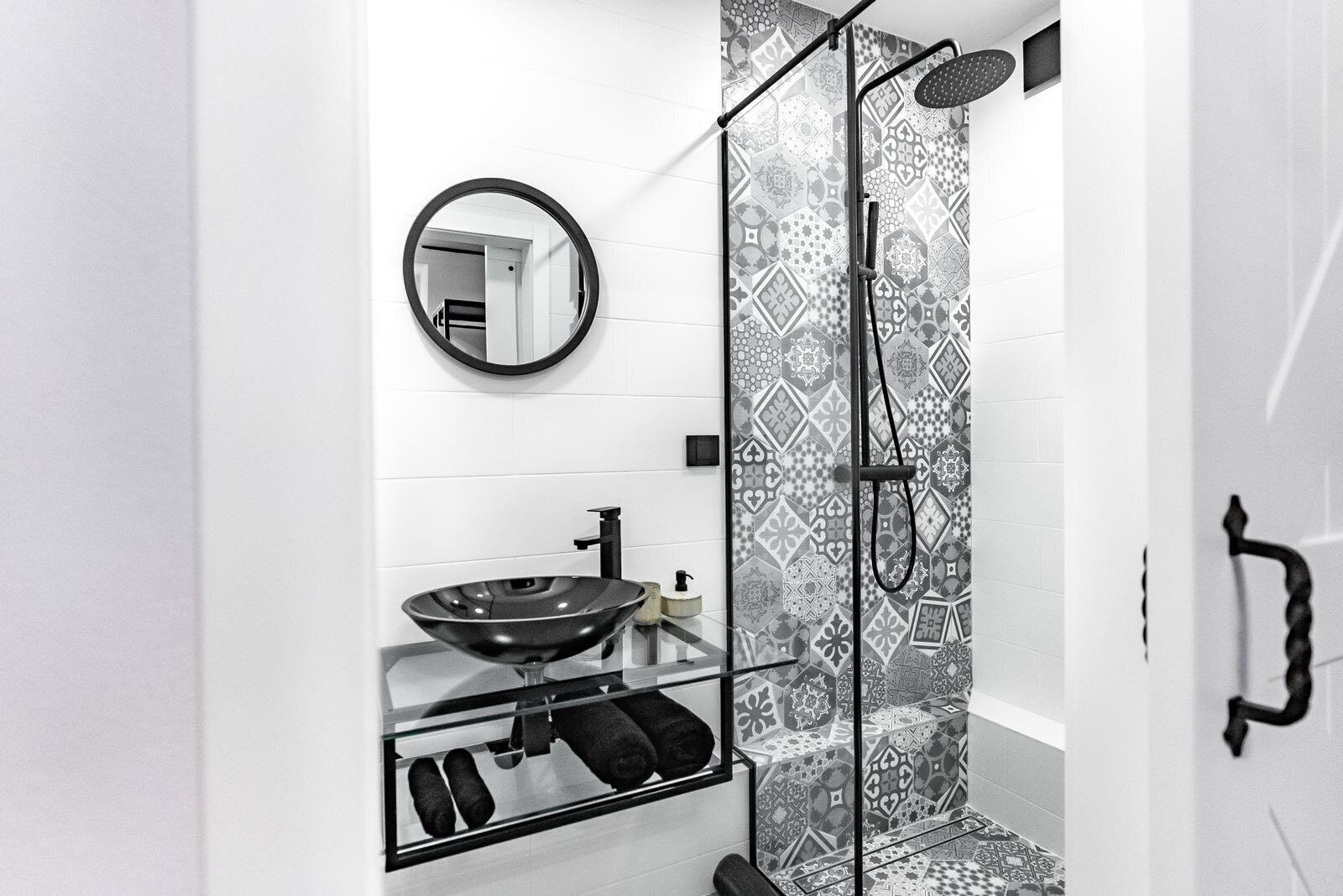 A modern bathroom with a black vessel sink, a round mirror, and a shower with patterned tiles.