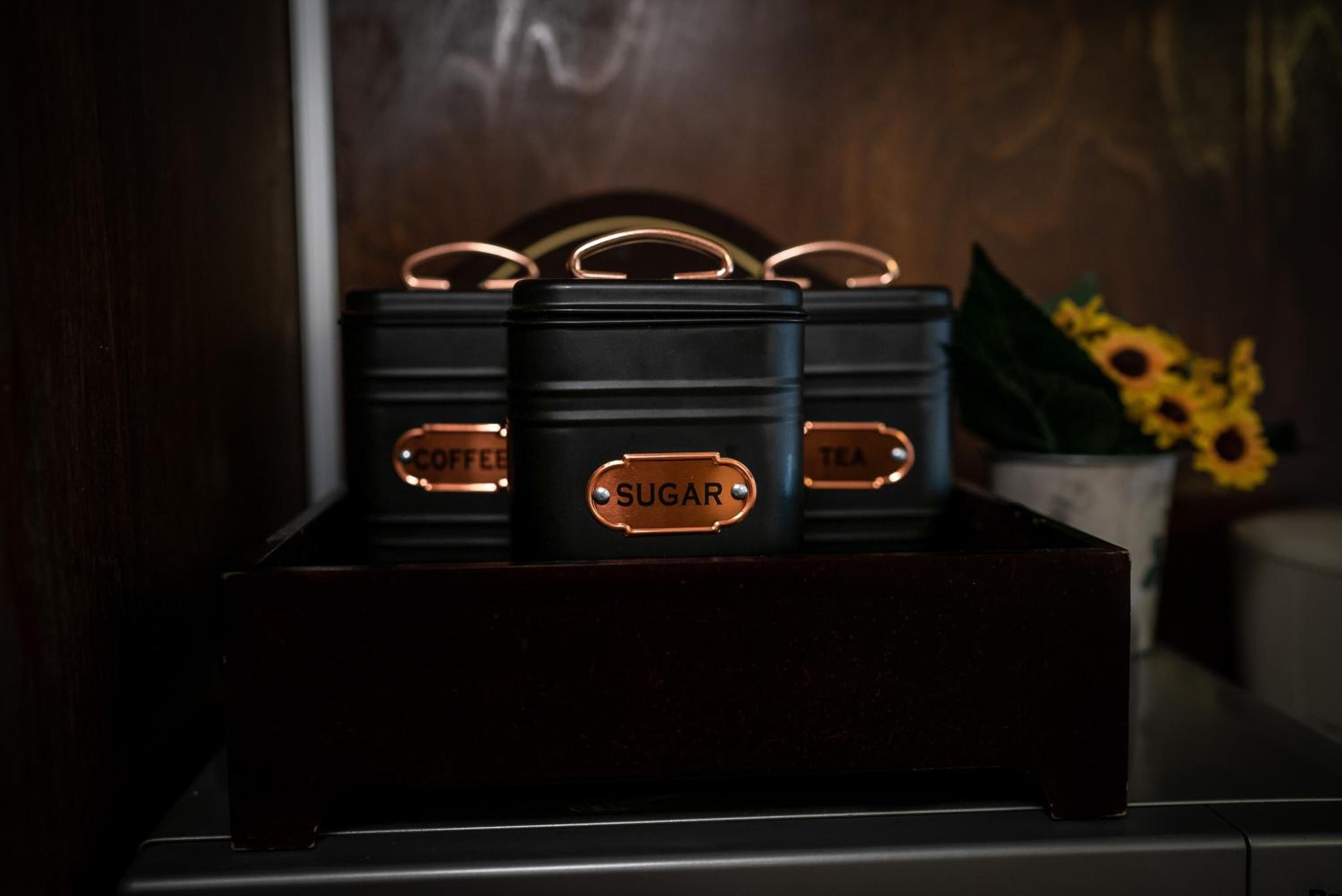 A set of black containers with copper handles and labels indicating 'COFFEE,' 'SUGAR,' and 'TEA' on a wooden surface.