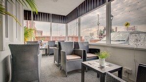 Un espacio exterior moderno con sillas de mimbre y una mesa, ventanas amplias con vista a la ciudad y una planta en maceta sobre la mesa.