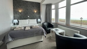 Habitación de hotel con cama, dos lámparas de noche, una silla, una mesita y vista a la playa a través de amplias ventanas.
