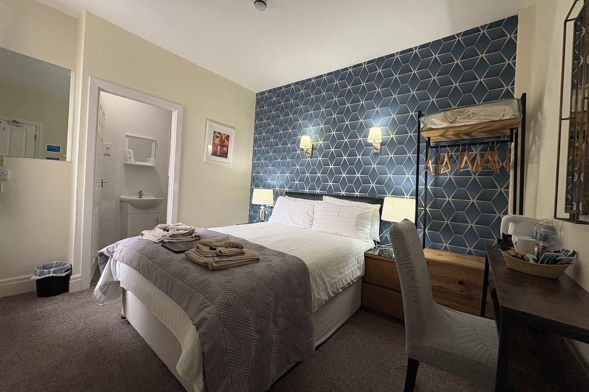 A hotel room with a bed, bedside tables, a desk, a chair, a mirror, a sink, and a patterned wall.
