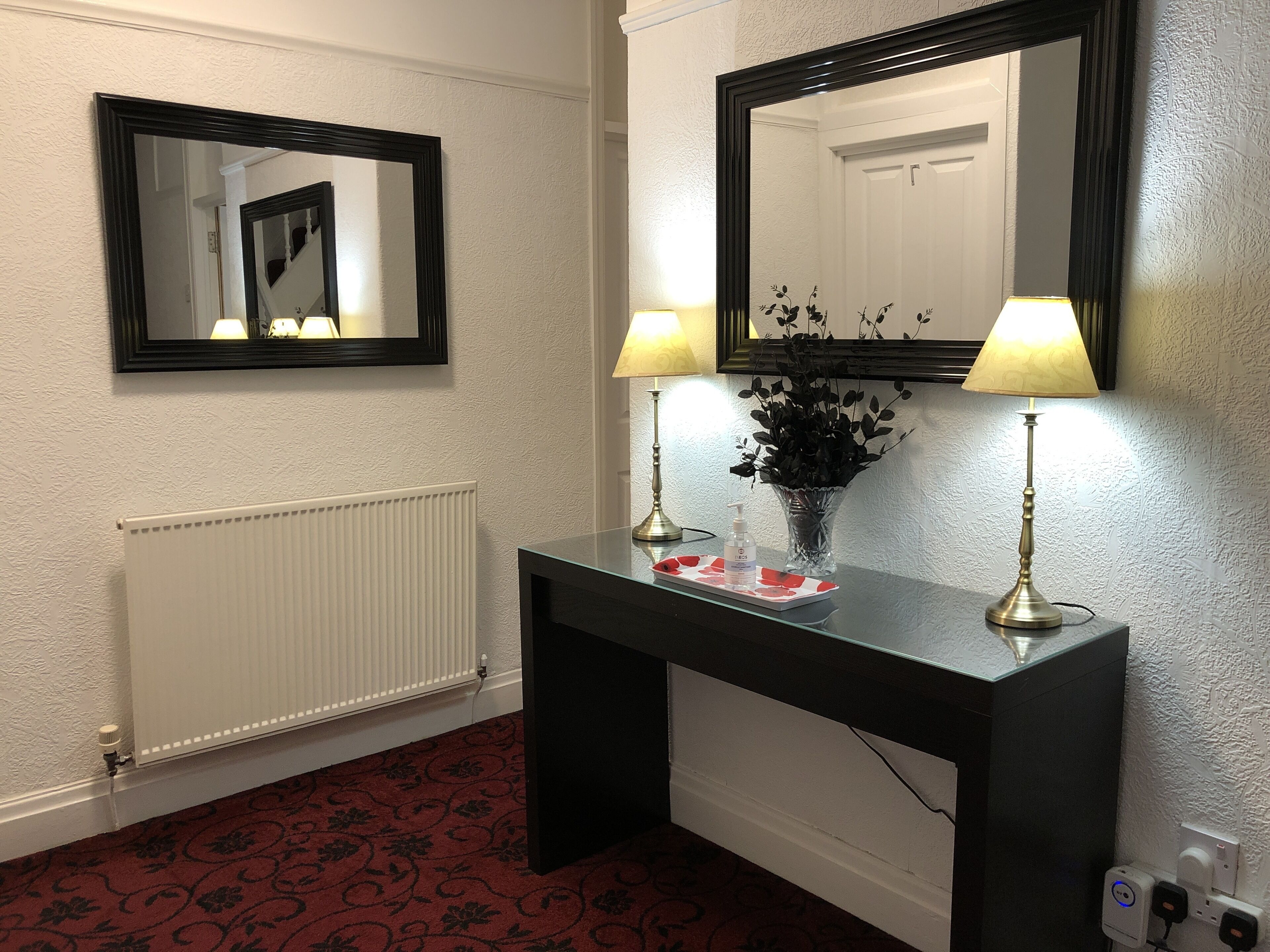 A hotel room with a black console table, two wall-mounted lamps, a mirror, a radiator, and a floral-patterned carpet.
