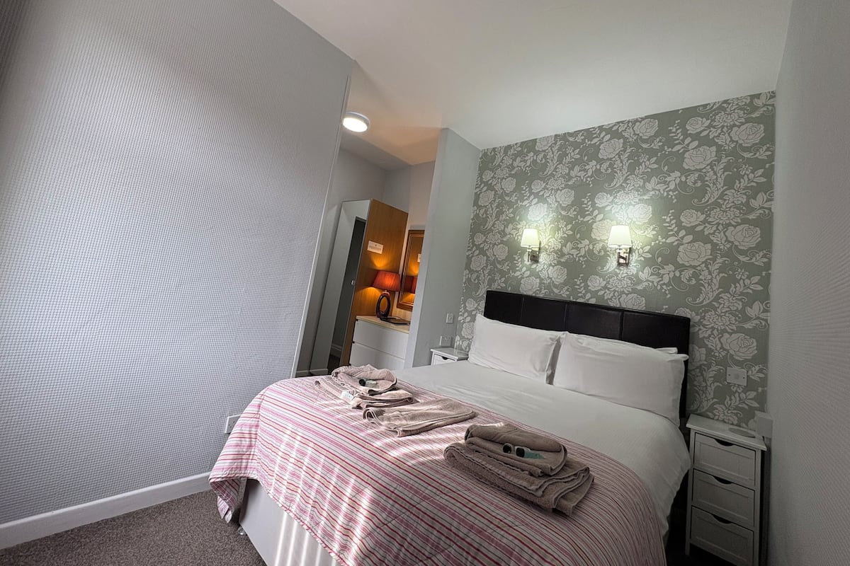 A hotel room with a bed, a nightstand, a mirror, and a wall with floral wallpaper.