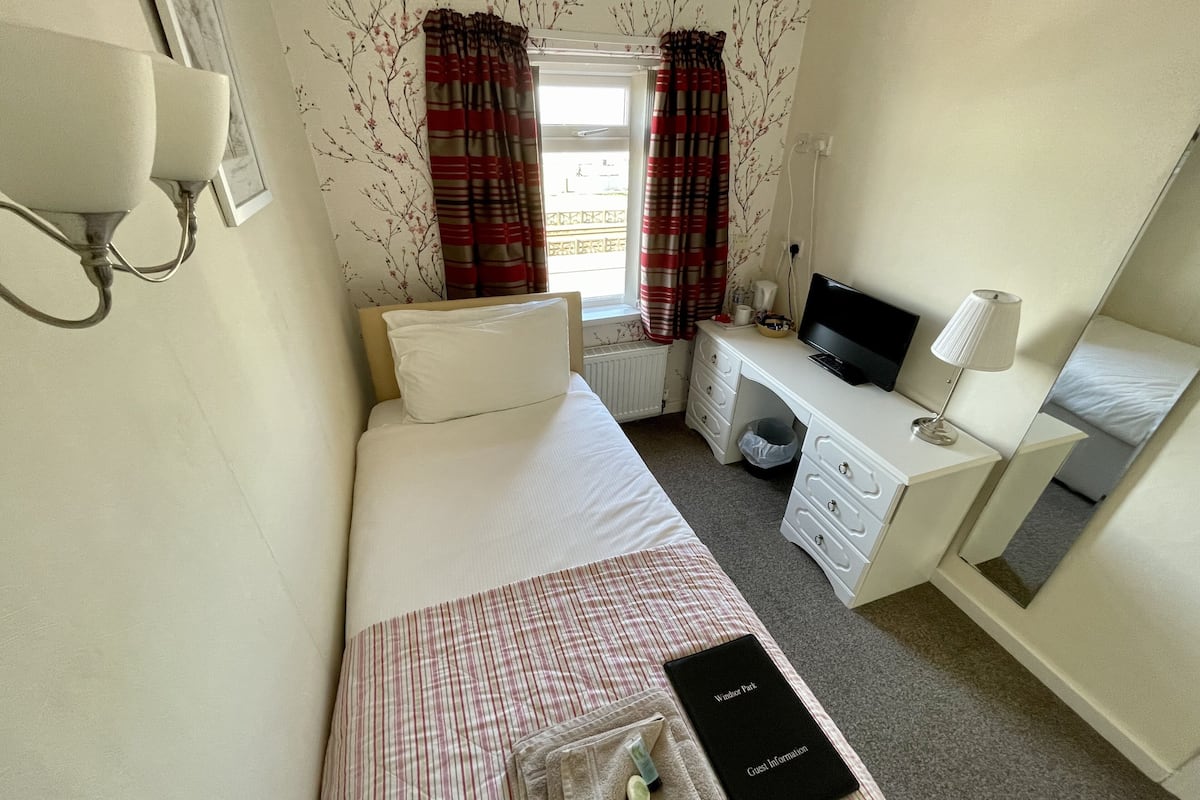 A hotel room with a bed, a desk, a chair, a TV, and a bedside table with a lamp.