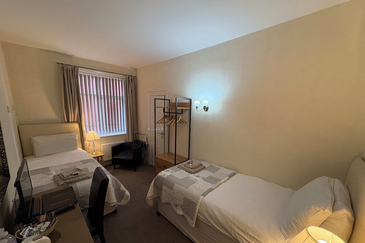 A hotel room with two beds, a desk, a chair, a coat rack, and a window with curtains.