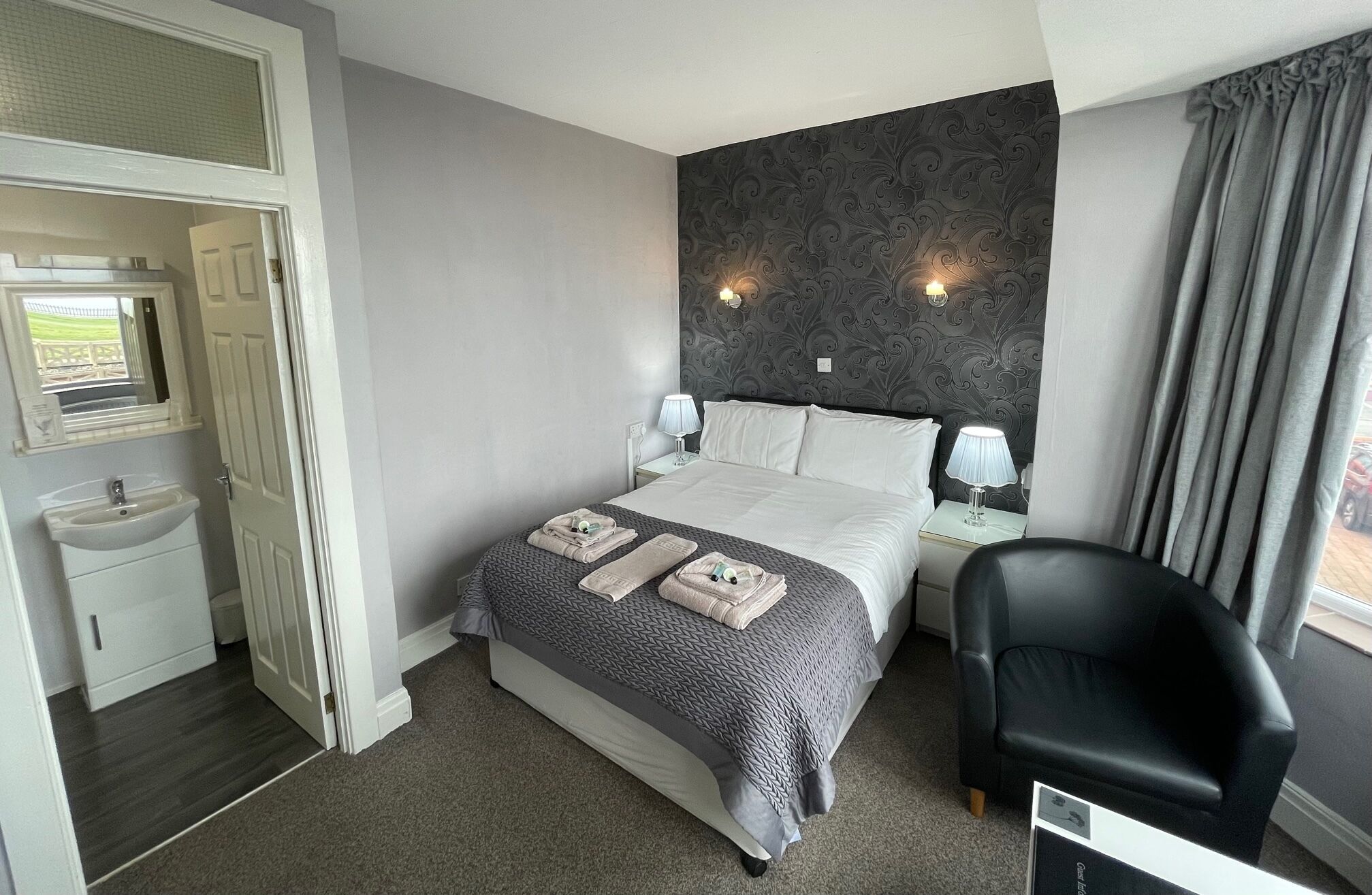 Een hotelkamer met een bed, bedlampen, een stoel, een kaptafel met spiegel en een raam met gordijnen.