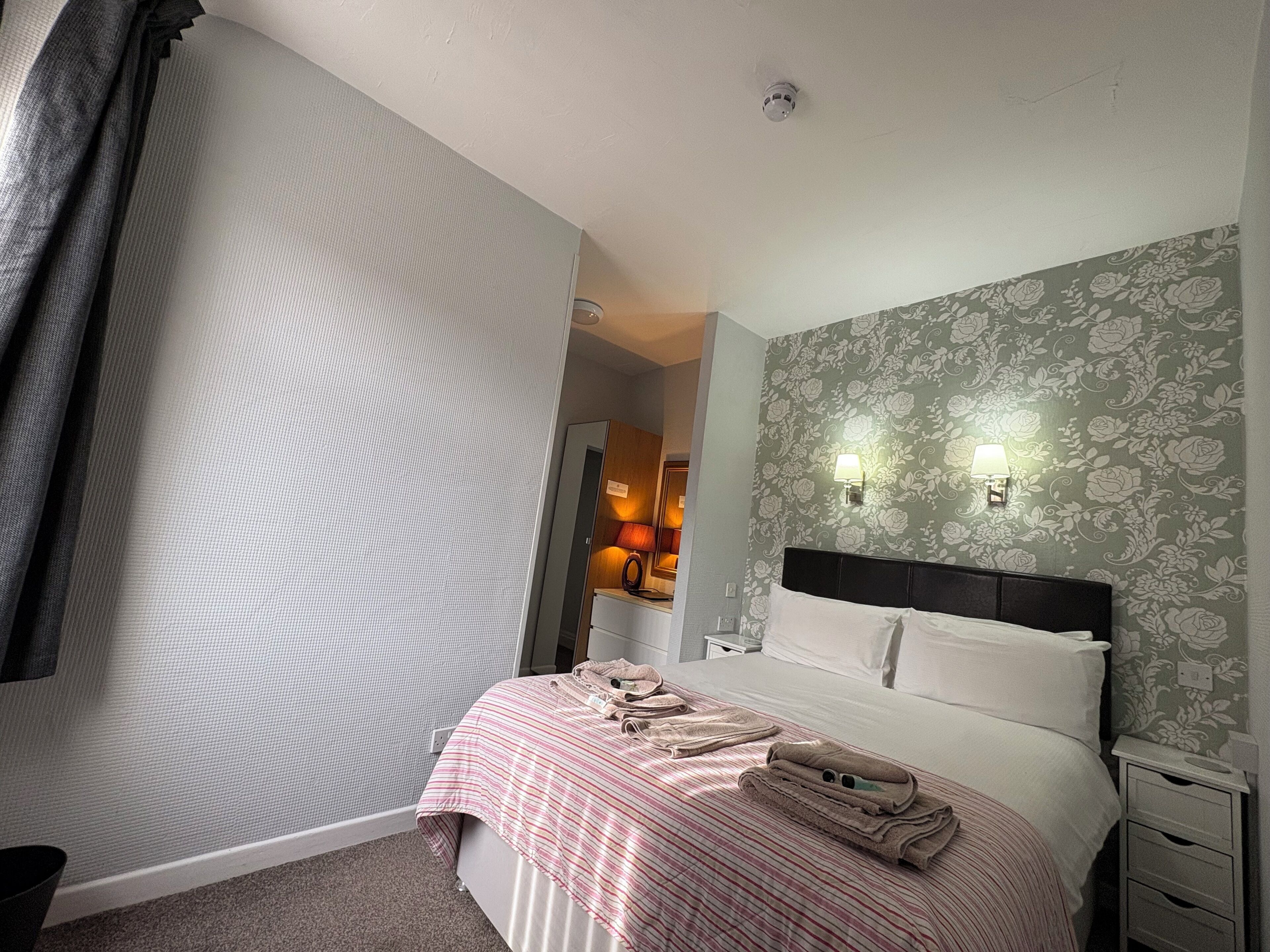 Habitación de hotel con cama, mesita de noche, espejo y baño visible a través de una puerta abierta.