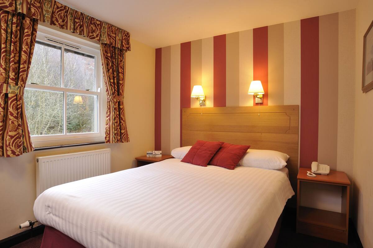 A hotel room with a large bed, two bedside lamps, a wooden headboard, a nightstand with a phone, and a window with curtains.