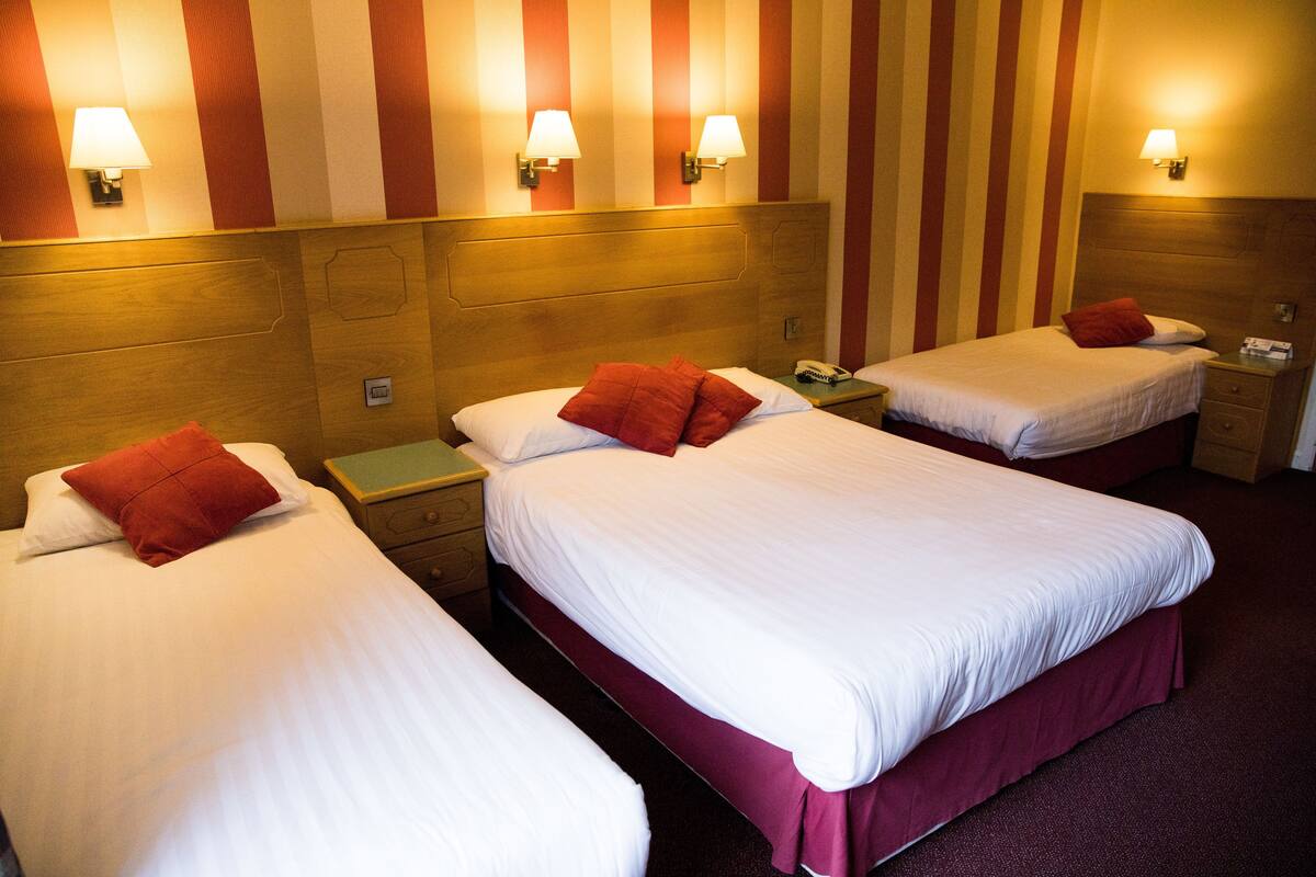 A hotel room with three beds, each with white bedding and red pillows. There are wall-mounted lamps on the wall above the beds.
