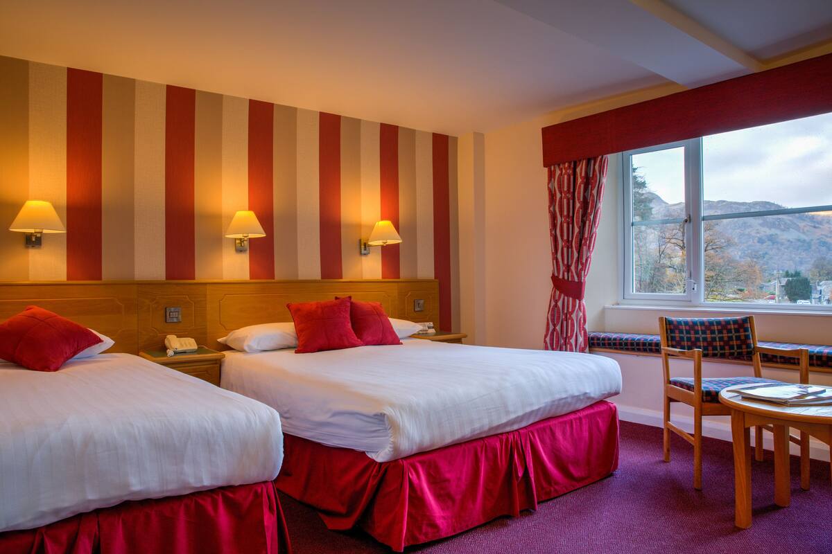 A hotel room with two beds, a window with a view of the mountains, a wooden chair, and a small table.
