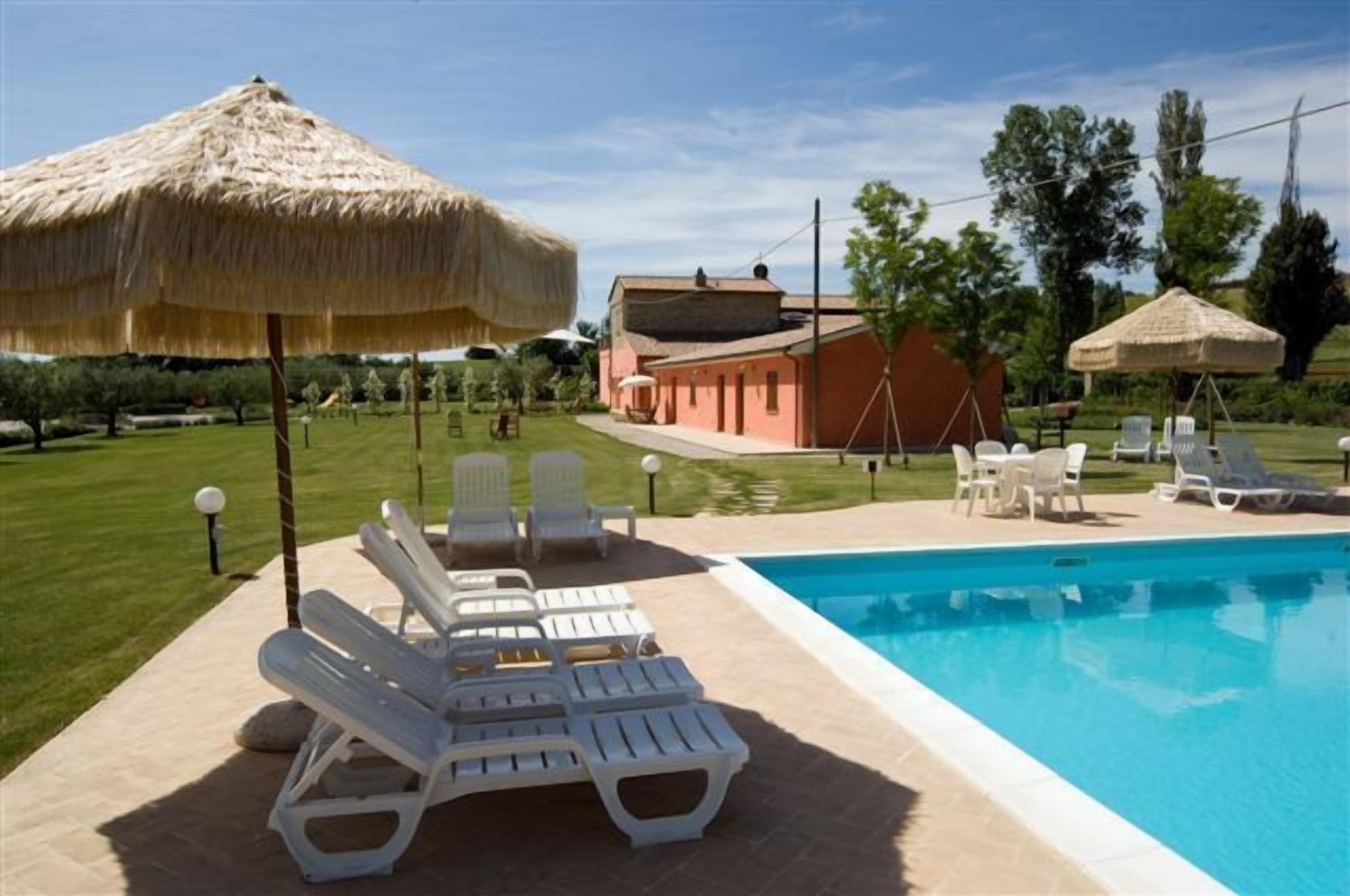 A swimming pool with lounge chairs and umbrellas, a building with a red roof, and a grassy area with trees.