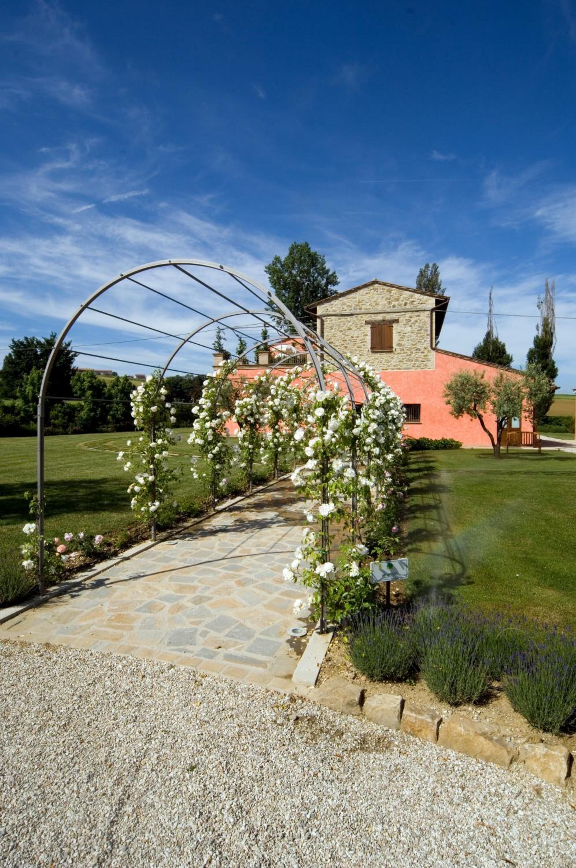 Un sentiero in pietra conduce a una casa con facciata in pietra e tetto rosso, circondata da un prato curato e piante fiorite.