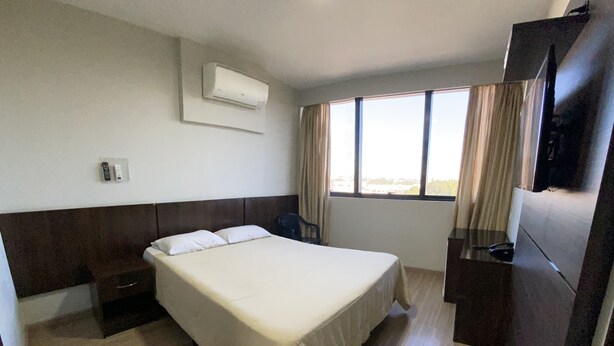Una habitación de hotel con cama, mesita de noche, escritorio, silla, televisor y ventana con cortinas.
