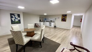 Amplia sala de estar con una mesa redonda de vidrio rodeada de sillas acolchadas, una cocina al fondo y obras de arte en las paredes.