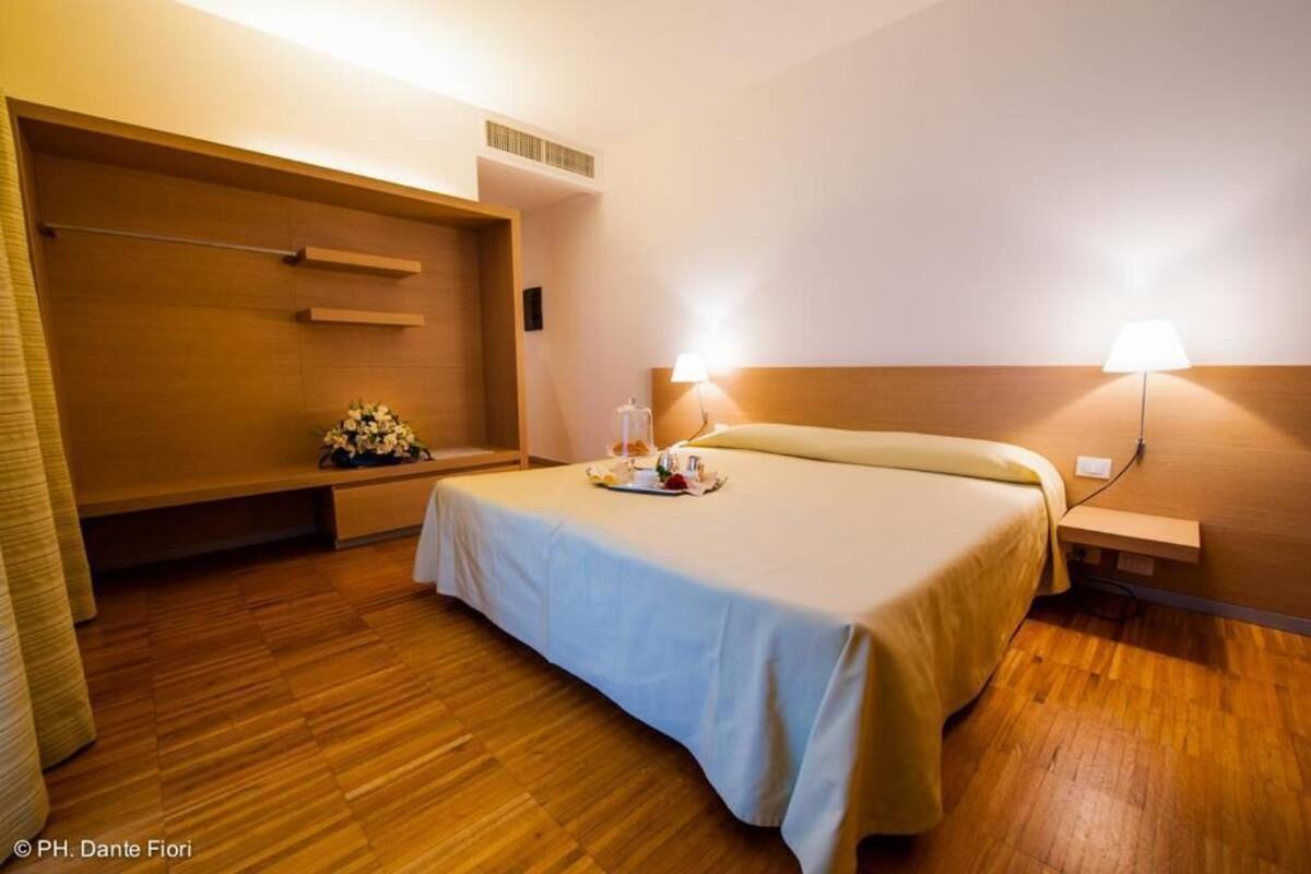 A hotel room with a bed, bedside lamps, a wooden headboard, a bench with a floral arrangement, and a tray with items on the bed.