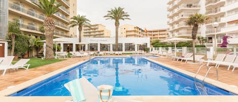 A swimming pool with lounge chairs and umbrellas, surrounded by palm trees and a hotel building.