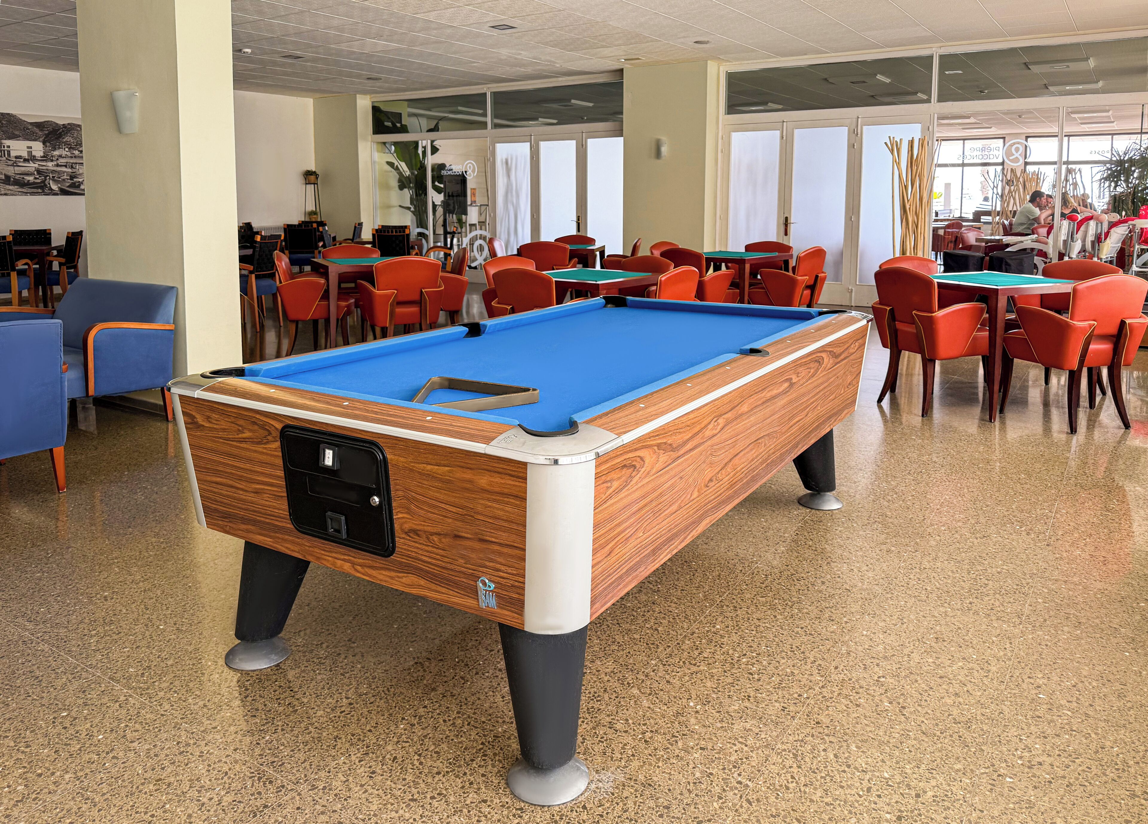 Ein Billardtisch in einem Raum mit Stühlen und Tischen, großen Fenstern und einer Decke mit eingelassener Beleuchtung.