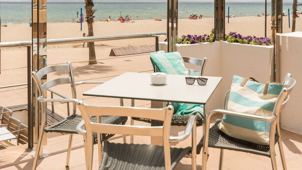 An outdoor patio with a table and chairs, overlooking a beach with palm trees and the sea. - Hotel Monterrey Roses by Pierre & Vacances (Roses)