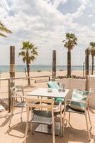 Eine Terrassensitzgelegenheit im Freien mit Tisch und Stühlen, mit Blick auf einen Strand mit Palmen und dem Meer.