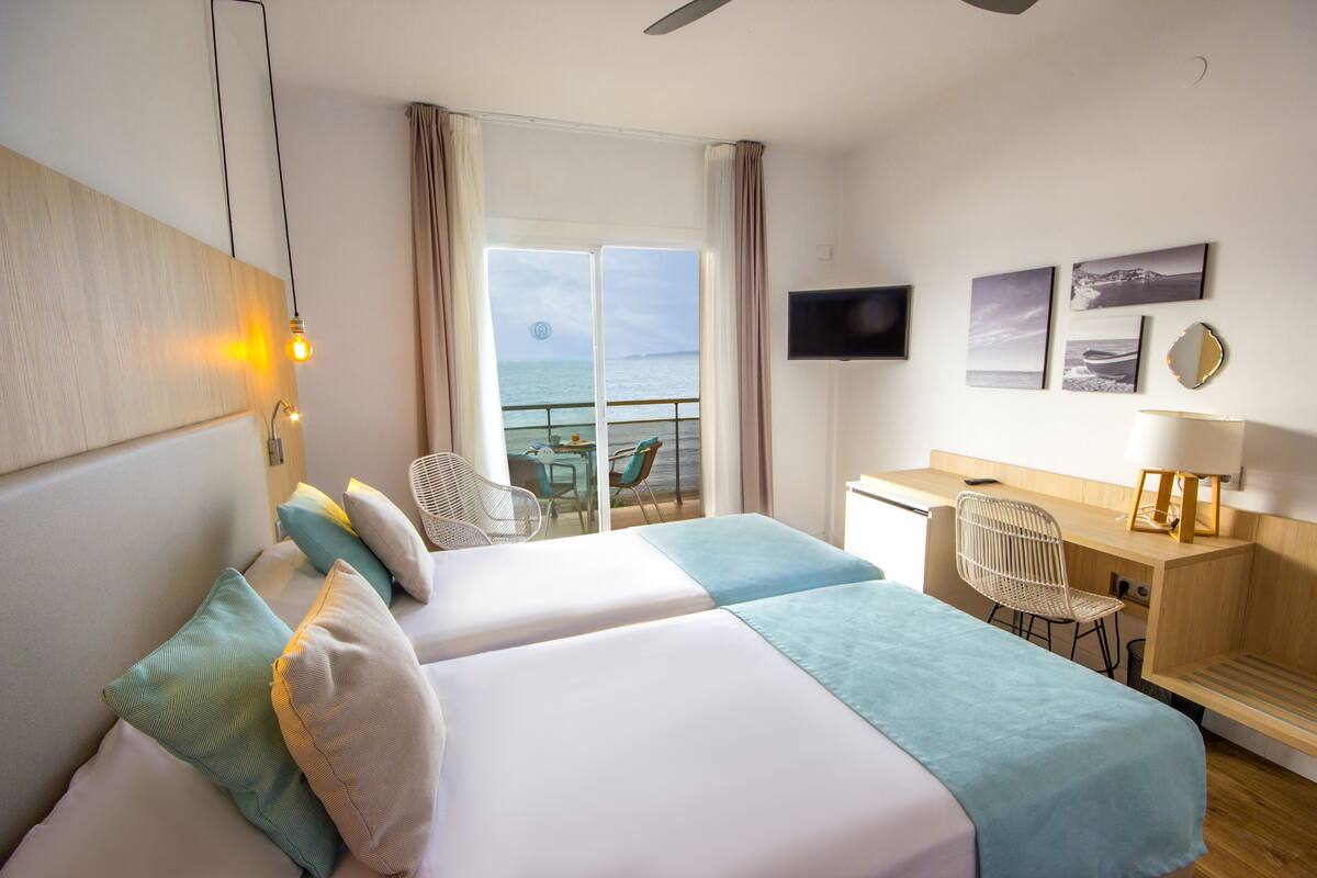 A hotel room with two beds, a desk, a chair, a TV, and a view of the sea.