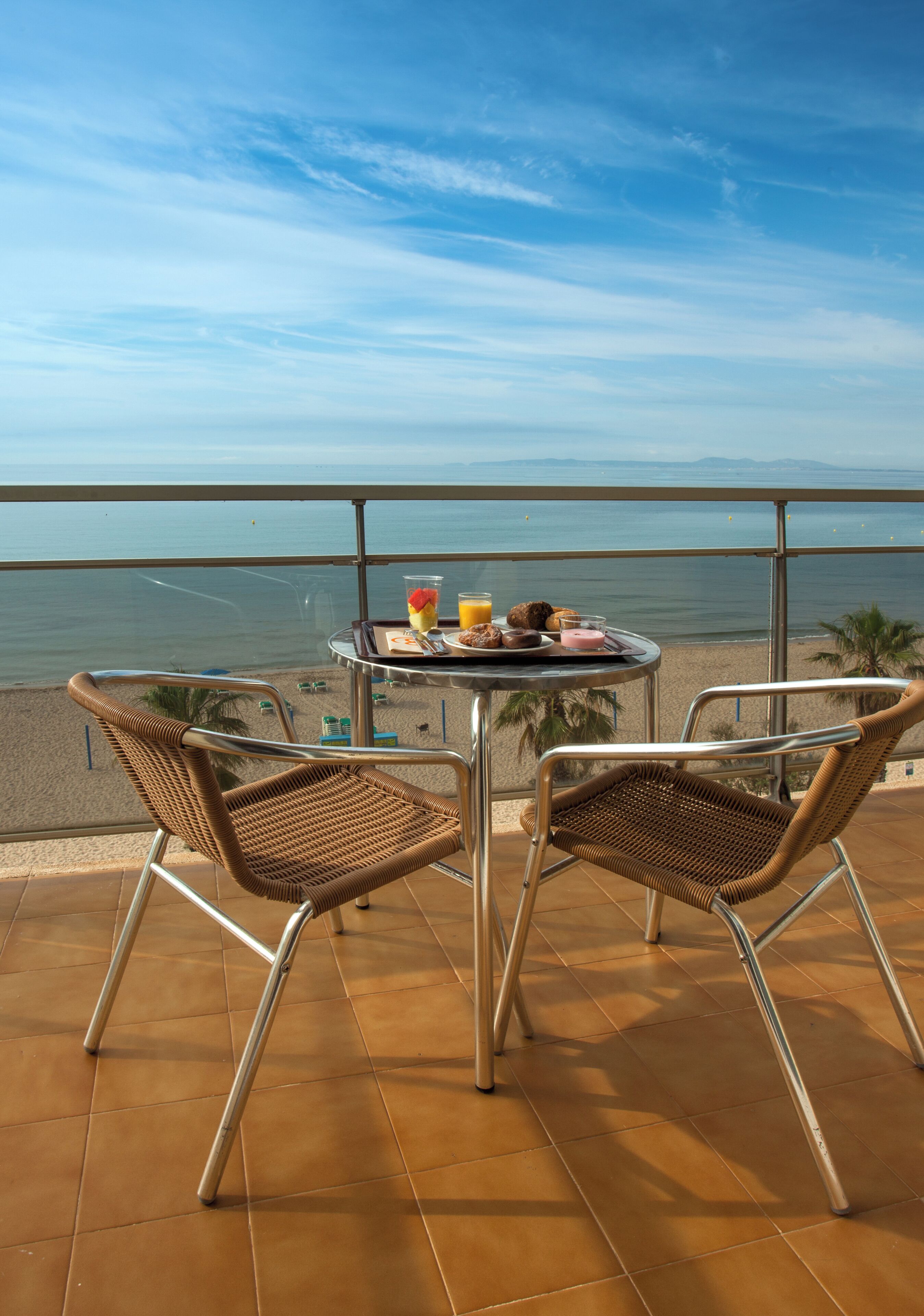 A balcony with two wicker chairs, a small round table with breakfast items, and a view of the sea.