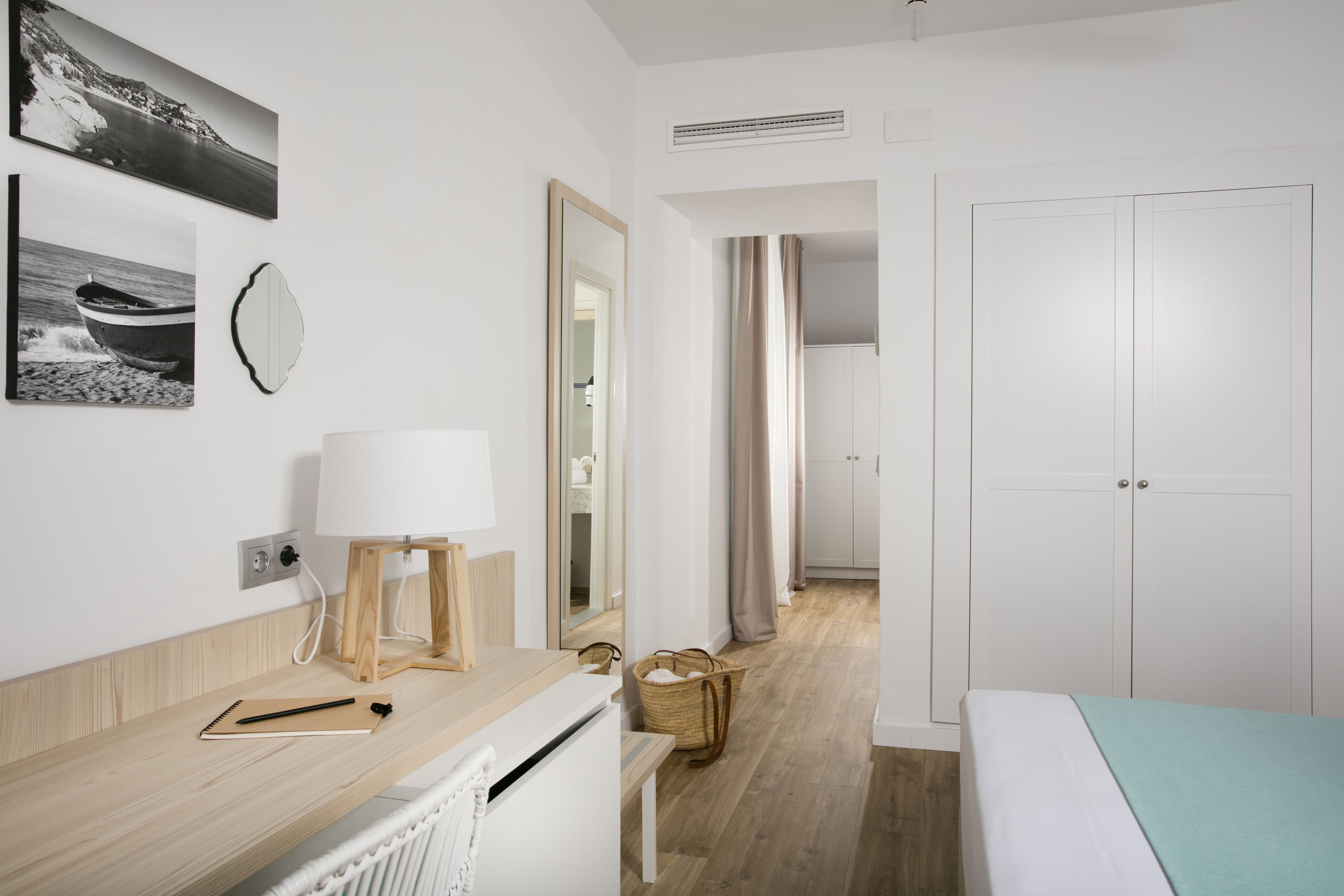Une chambre d’hôtel moderne avec un bureau en bois, un lit recouvert d’une couverture bleu clair, un radiateur blanc, un miroir et une vue sur le couloir.