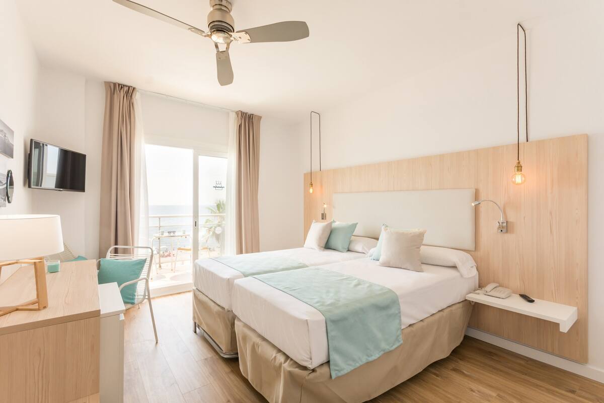 A hotel room with two beds, a desk, a chair, a TV, and a balcony with curtains.
