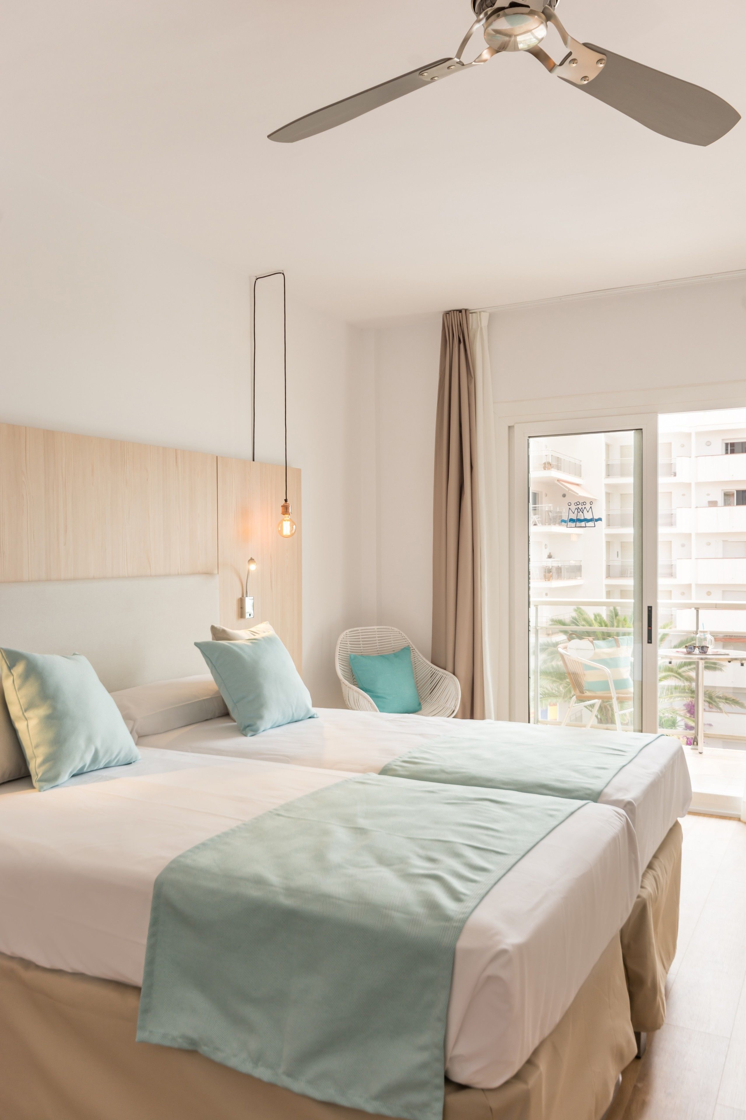 A hotel room with a bed, a ceiling fan, a chair, and a balcony with a view of buildings.