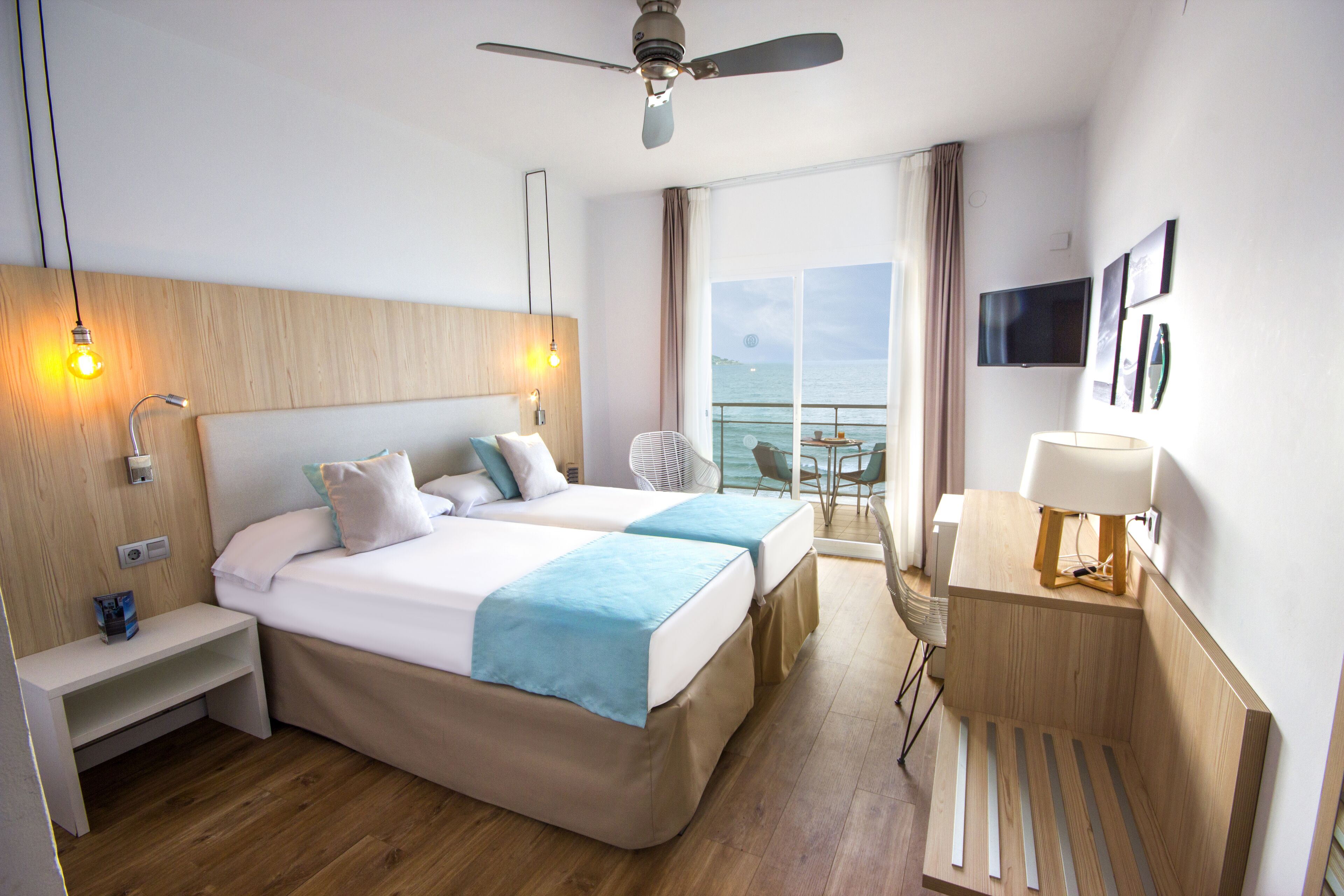 A hotel room with two beds, a desk, a chair, a TV, and a balcony with a view of the sea.