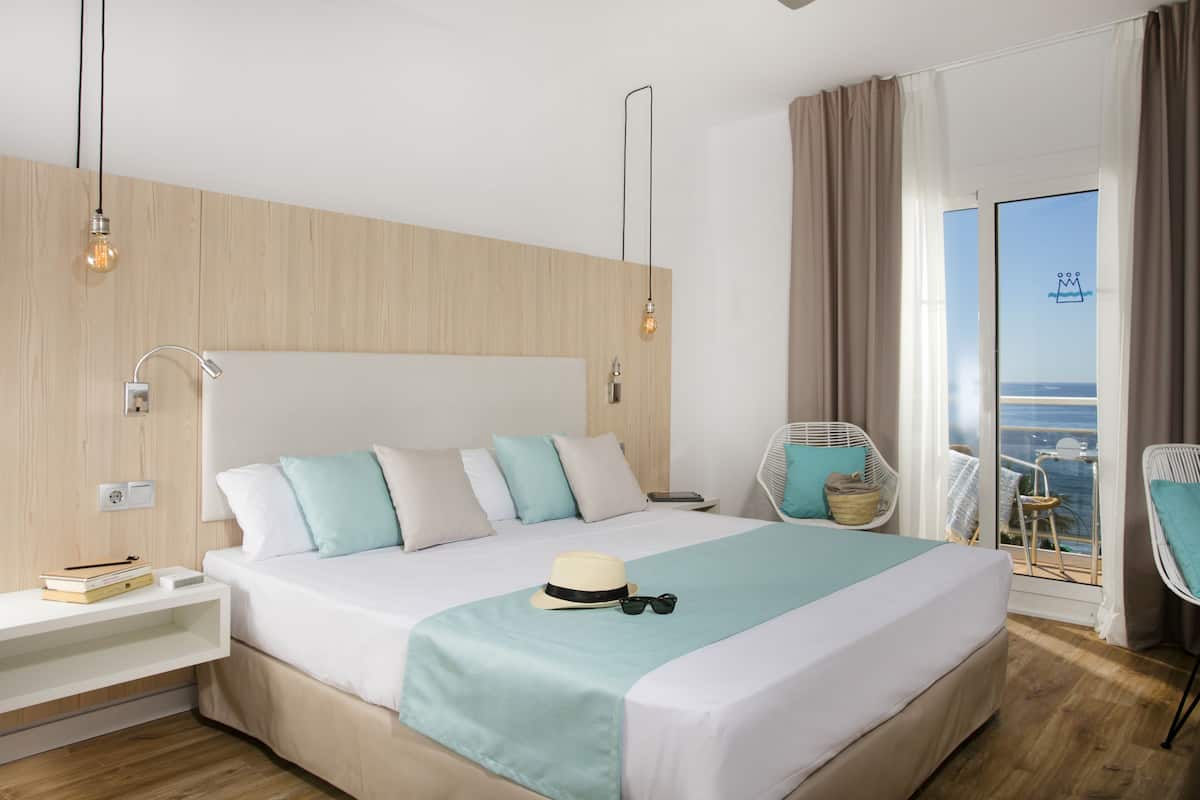 A modern hotel room with a large bed, bedside tables, a view of the sea, and a balcony with a chair.
