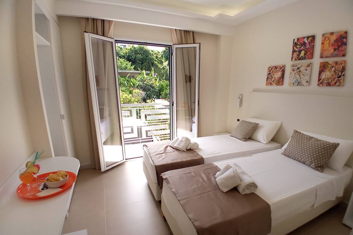 A hotel room with two beds, a dining table with a tray of food, and a balcony with a view of greenery.