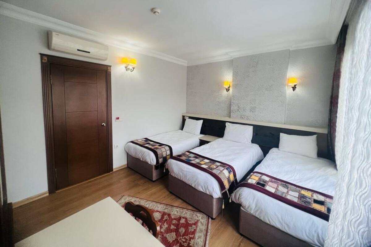 A hotel room with three single beds, a wooden wardrobe, a desk, and two wall-mounted lamps.