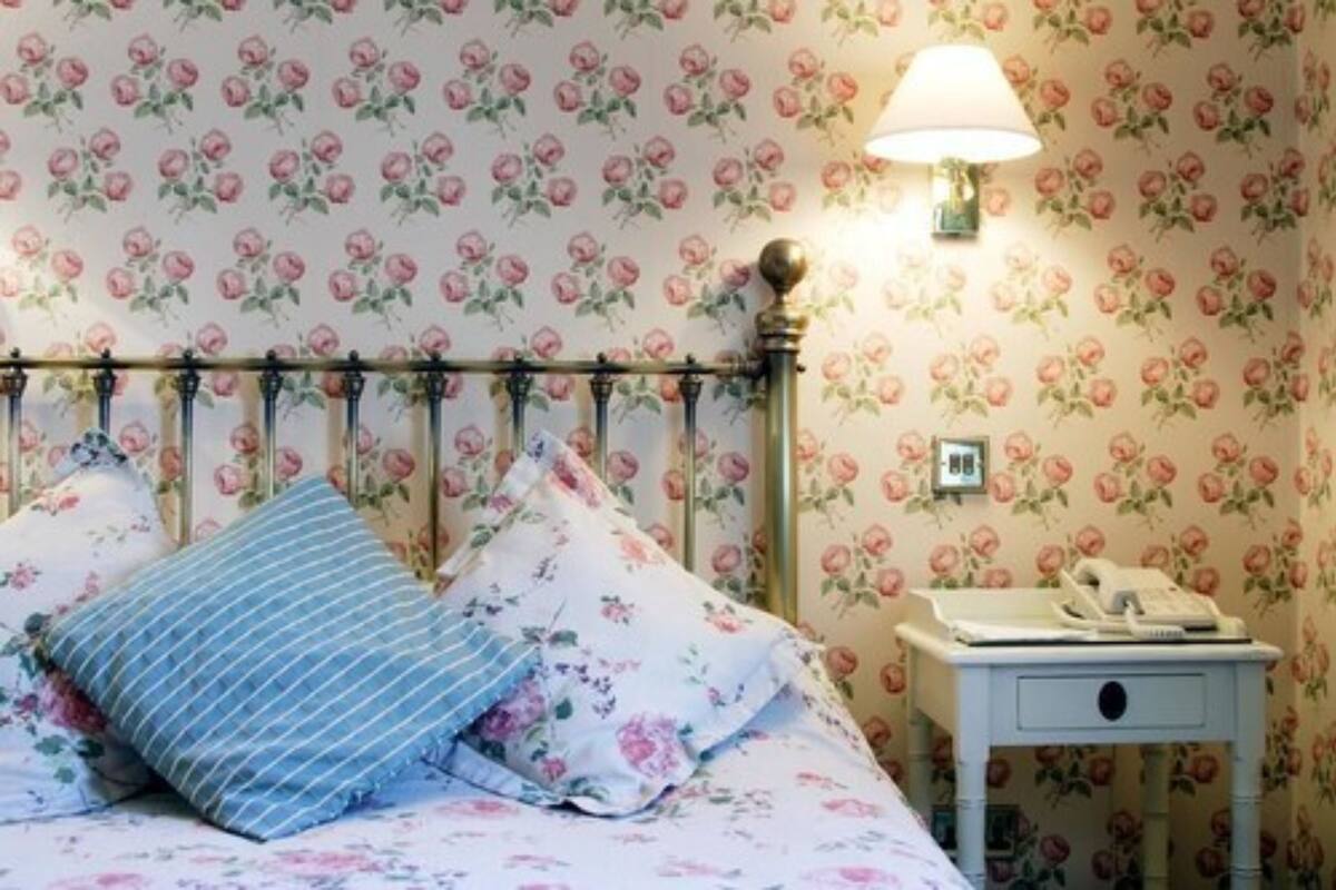 A bedroom with floral wallpaper, a bed with floral bedding, a bedside lamp, and a nightstand with a telephone.