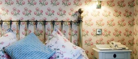 A bedroom with floral wallpaper, a bed with floral bedding, a bedside lamp, and a nightstand with a telephone.
