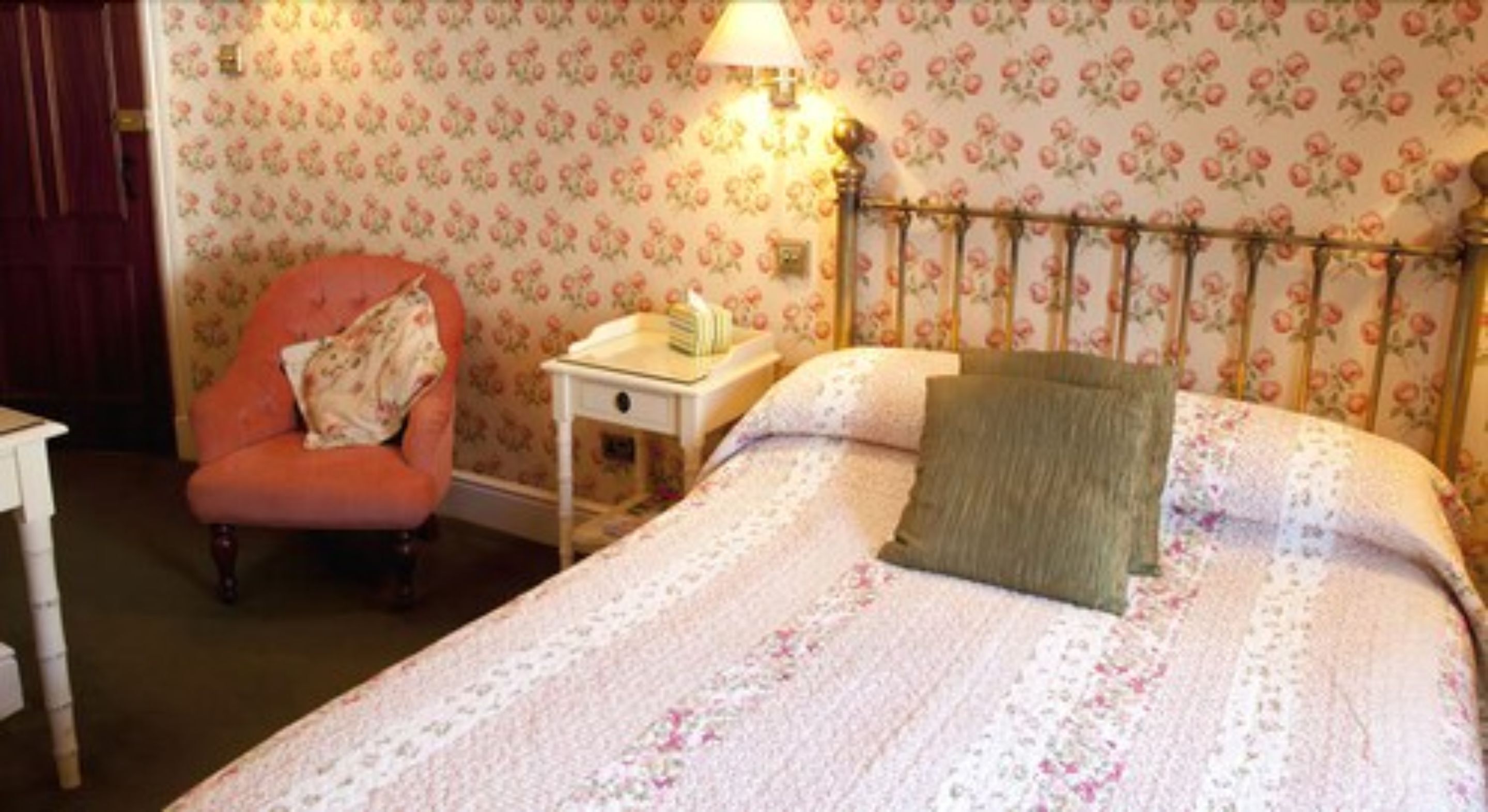 A bedroom with a floral wallpaper, a bed with a green pillow, a nightstand, and a chair.