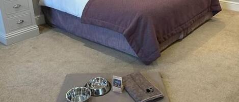A bedroom with a tufted headboard, a bed with a purple bedspread, a bedside table with a lamp, and a small rug with pet bowls.