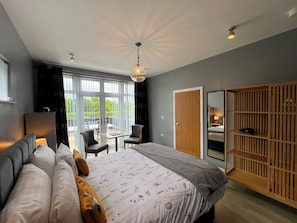 A modern bedroom with a large bed, a wooden wardrobe, a desk, and a balcony with a view.