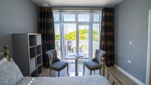 Un dormitorio con cama, escritorio, dos sillas, estantería y vistas al exterior.
